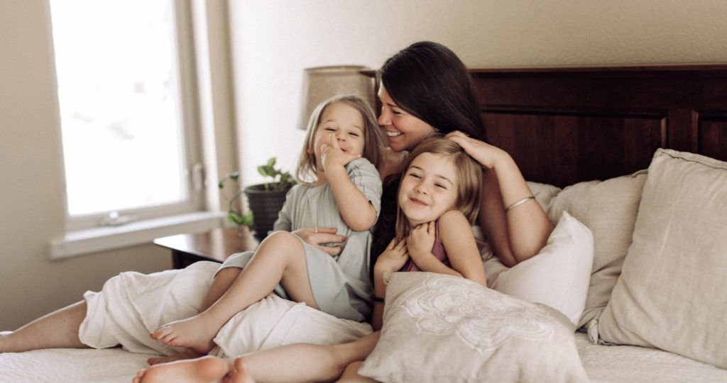 A mother laughing with her two young daughters on a sunlit bed